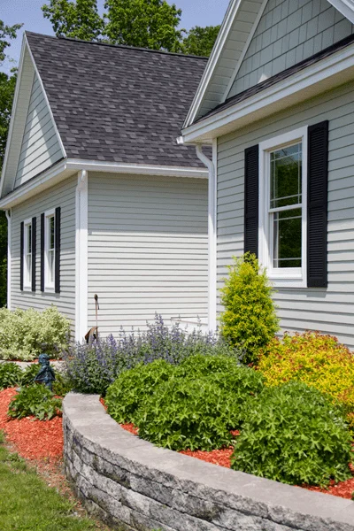 Corner of a gray-green Vermont home with white seamless gutters above a landscaped stone retaining wall